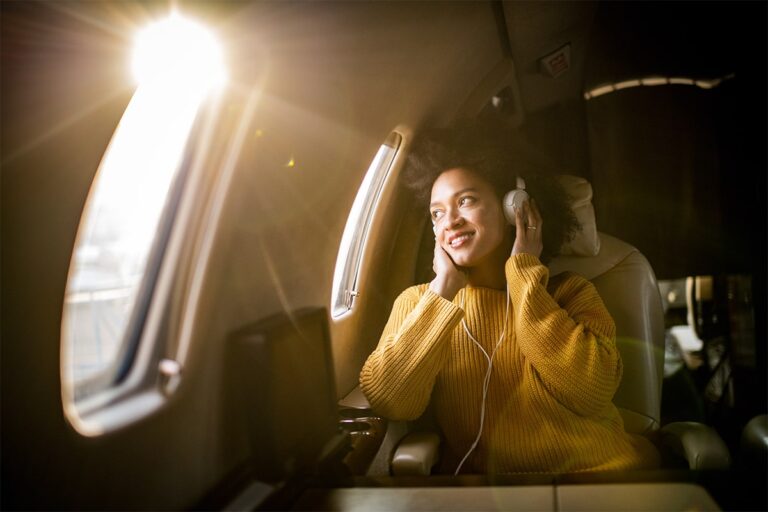 A young woman on an airplane looking out the window with headphones on her ears.