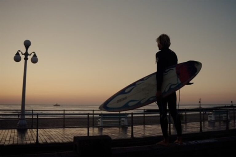 A surfer standing on the boardwalk looking out to the ocean while holding a surfboard at sunset.